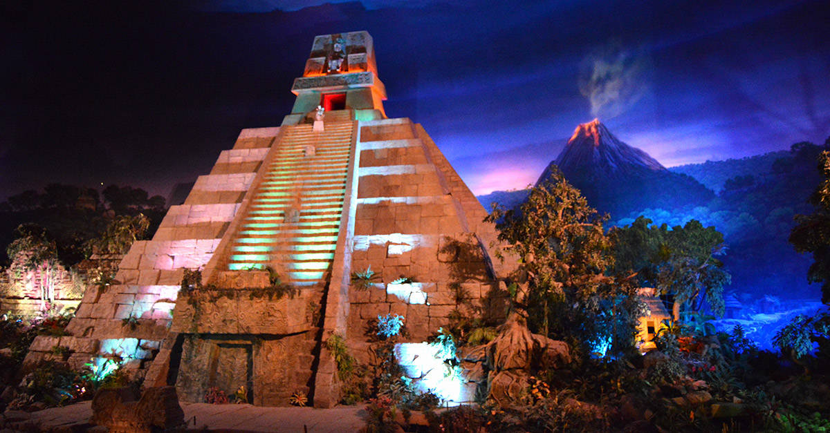 A glowing ancient pyramid creates a dazzling nighttime spectacle, set before a dramatic volcano in this thrilling themed adventure.