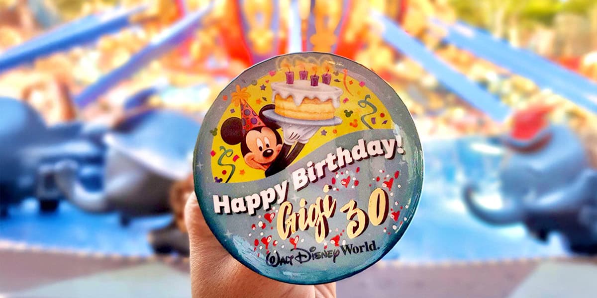 A hand holds a Disney World birthday button featuring Mickey Mouse, a cake with candles, and the words "Happy Birthday! Greg 30"—a perfect photo for anyone seeking creative Disney trip ideas, with the colorful Dumbo ride in the background.