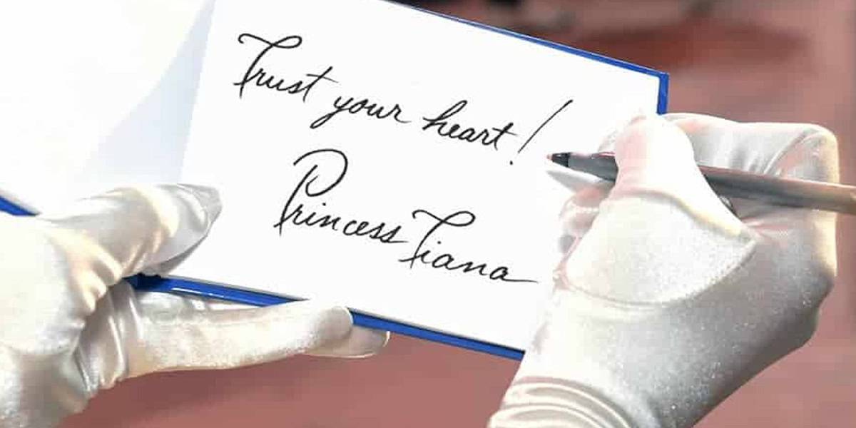 A person wearing white gloves writes "Trust your heart! Princess Diana" in cursive on an open card with a pen, inspired by creative Disney trip ideas.