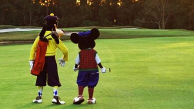 Two people in Goofy and Mickey Mouse costumes, dressed in vintage golf outfits, walk side by side on a golf course, with Goofy carrying a golf bag and Mickey holding a golf club. Trees and sand traps are in the background.