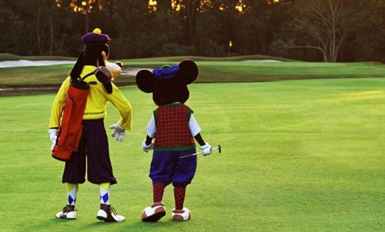 Two people in Goofy and Mickey Mouse costumes, dressed in vintage golf outfits, walk side by side on a golf course, with Goofy carrying a golf bag and Mickey holding a golf club. Trees and sand traps are in the background.