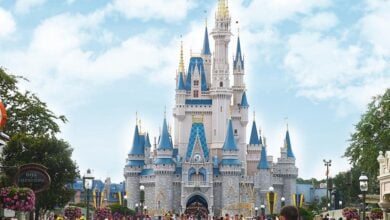 A crowd of people gathers in front of a large, ornate castle with blue and gold spires at a theme park, surrounded by trees and flowers under a bright, partly cloudy sky.