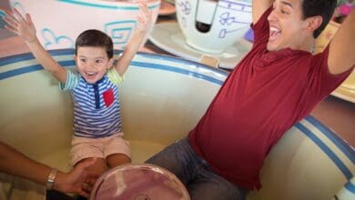 A young boy and a man smile and raise their arms in excitement while riding together in a large spinning teacup at an amusement park—one of many magical moments you’ll find with the right Disney Park Tips.