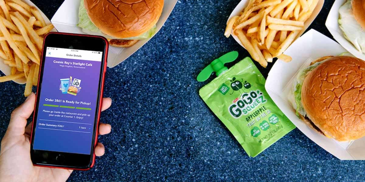 A person holds a phone displaying a food order screen, like you’d see in the Disney Pro Guide, next to trays with burgers, fries, and a green applesauce pouch on a blue table.