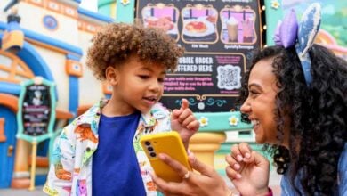 A smiling woman with mouse ears, looking like a true Disney Pro Guide, holds a yellow phone and shows it to a young boy in a colorful shirt. They are outdoors, in front of a whimsical menu sign at an amusement park.