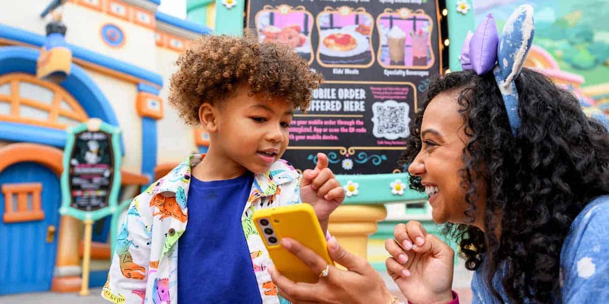 A smiling woman with mouse ears, looking like a true Disney Pro Guide, holds a yellow phone and shows it to a young boy in a colorful shirt. They are outdoors, in front of a whimsical menu sign at an amusement park.