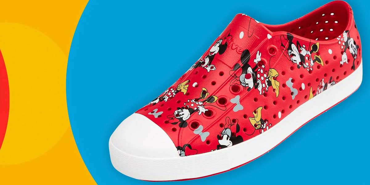 A red slip-on shoe with a white toe cap, decorated with Disney Pro Guide characters like Mickey Mouse and Minnie Mouse, and small white polka dots. The background features a bright blue and yellow design.