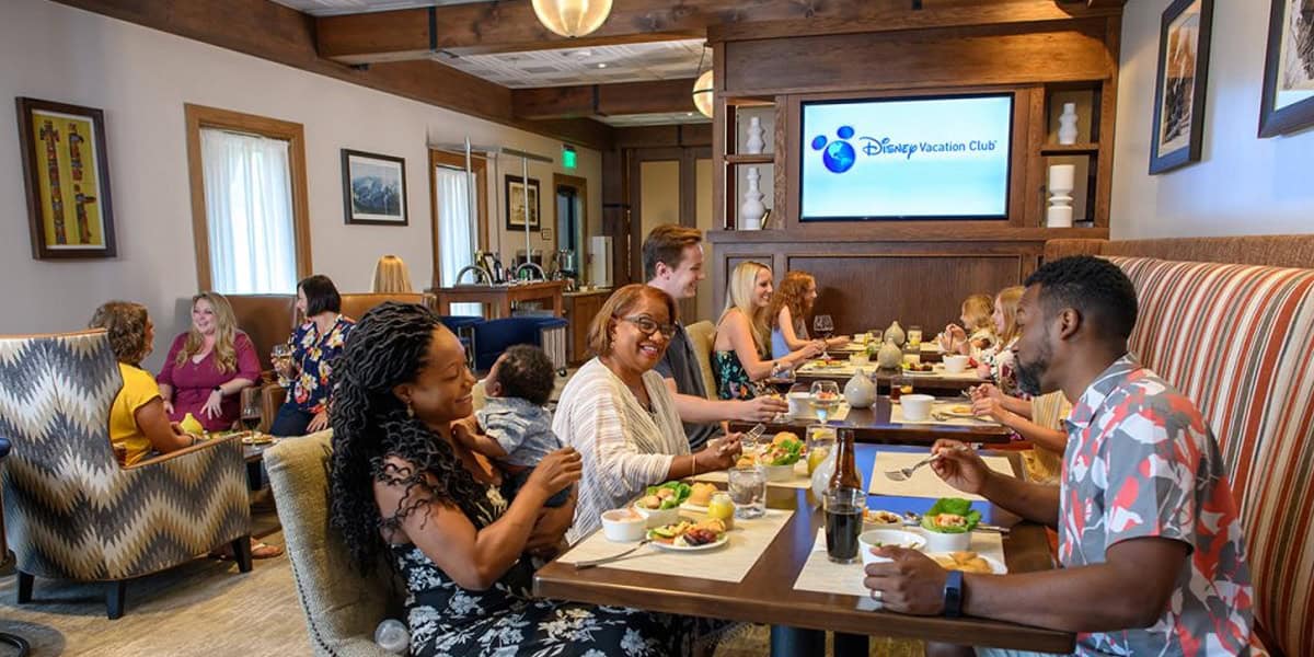 People of different ages and backgrounds dine together at a lively restaurant, smiling and talking over food. The atmosphere is warm and inviting, with wooden décor and a Disney’s Wilderness Lodge sign on the wall.