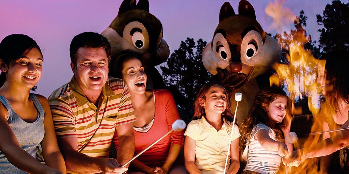 A family roasts marshmallows over a campfire at dusk, smiling together. Two people in large chipmunk costumes stand behind them, adding Disney Nighttime Experiences magic. Trees and a purple sky are visible in the background.