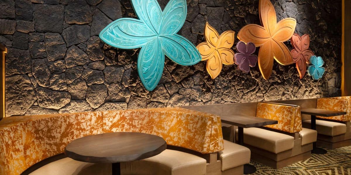 A cozy seating area with cushioned booths against a stone wall. Large, colorful floral sculptures in blue, orange, and purple are mounted on the wall, adding a vibrant touch to the dimly lit space. Round wooden tables complement the setup.