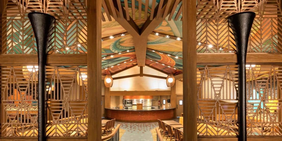 The image shows a warm, inviting dining area with wooden accents and artistic designs on the ceiling. The space features symmetrical decor, chairs, tables, and ambient lighting, creating a cozy atmosphere.