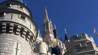 Close-up view of a castle with light stone walls, blue conical roofs, and golden spires against a clear blue sky. The image captures intricate architectural details.