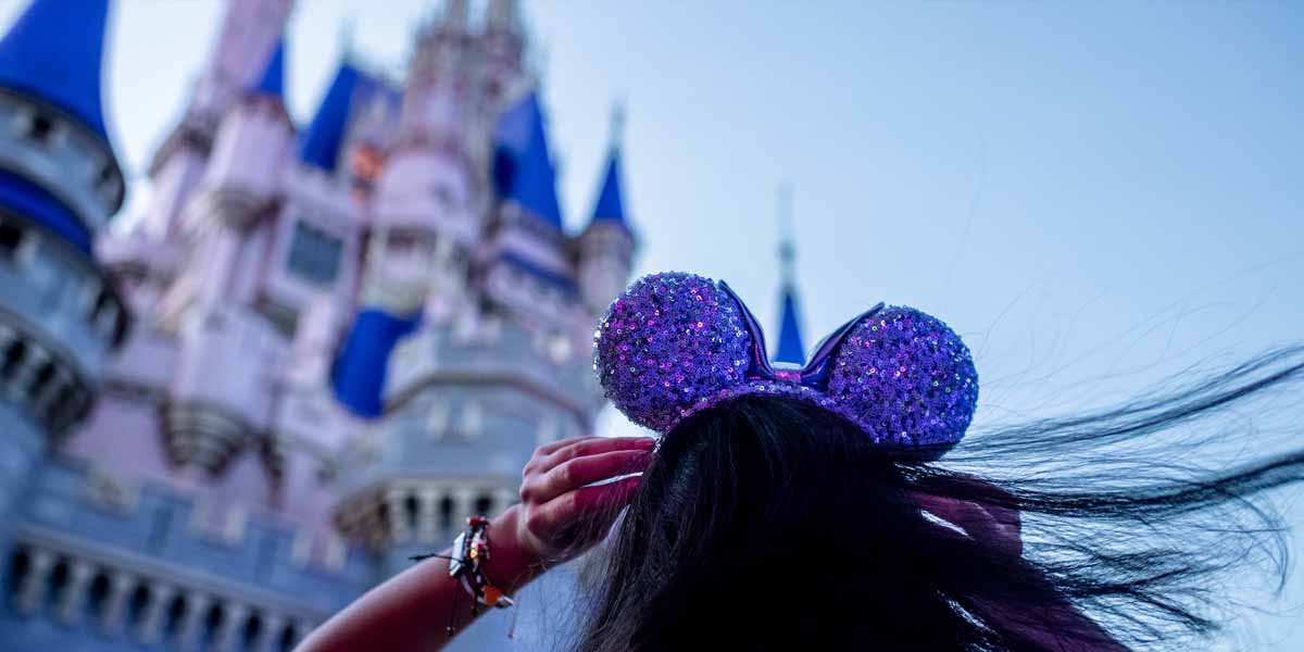 A person with long hair wearing sparkly purple Mickey Mouse ears stands in front of a pink and blue fairytale castle, likely at a theme park, looking ready to save money at Disney World with their hands touching the headband.