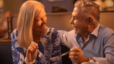 An older couple sits together in a restaurant booth, smiling and laughing while holding forks, enjoying delicious Epcot meals and each other’s company.