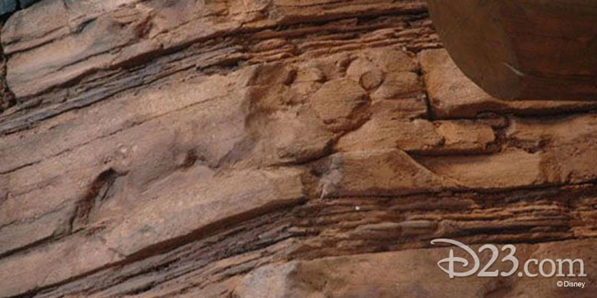 A close-up of textured brown rock layers with a hidden Mickey Mouse shape subtly formed in the stone. Discover fun Hidden Mickey facts while spotting the D23.com and Disney logos in the corner.
