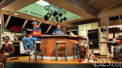 A kitchen scene features animated human figures in 1990s attire using various gadgets, a dog with a red bow sits in front, and a woman works at a computer; the room, filled with Tomorrowland Secrets, is decorated for Christmas.