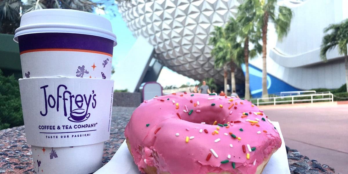 A Joffrey’s coffee cup and a pink frosted donut with colorful sprinkles sit on a ledge, captured near one of the Disney World Food Carts, with palm trees and Epcot’s Spaceship Earth geodesic sphere in the background.