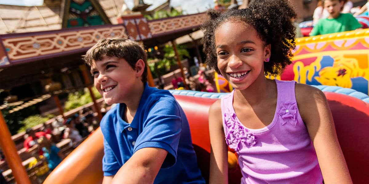 Two smiling children, a boy in a blue shirt and a girl in a pink top, sit together on a colorful amusement park ride with cheerful expressions—making memories and enjoying the day with Disney World tips for families in the background bustle.