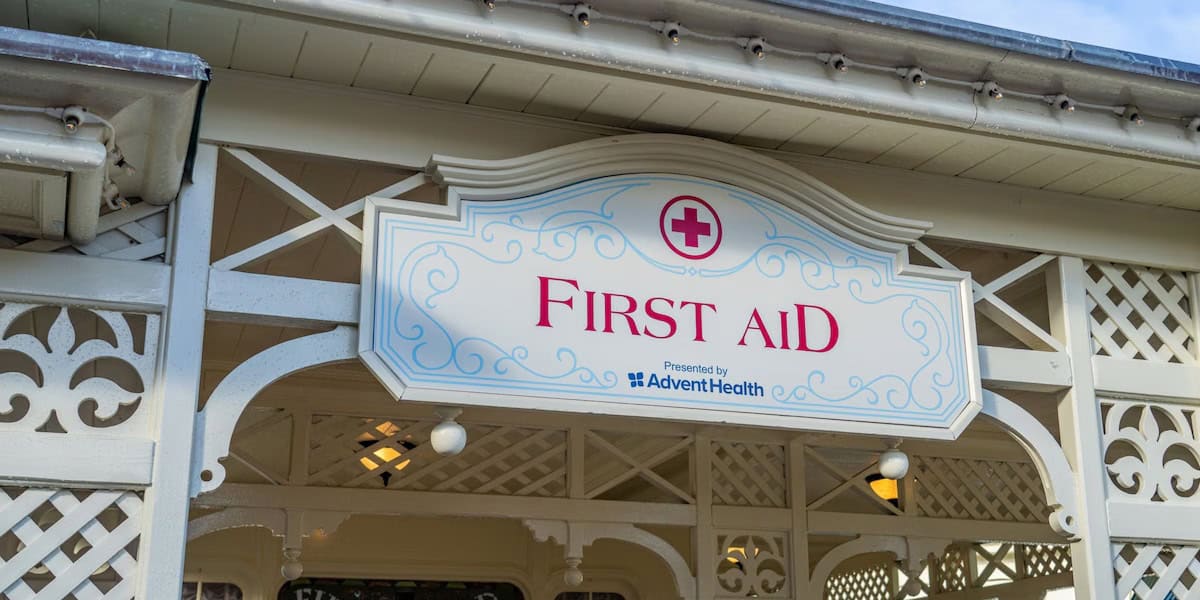 A white sign with red letters reading "First Aid" and a red cross symbol hangs above a decorative wooden structure, promoting Disney World Safety, with "Presented by AdventHealth" written below.