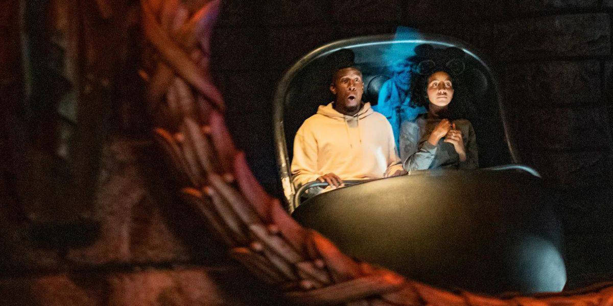 Two people sit together in a dark ride vehicle, both looking ahead with surprised expressions. The dimly lit scene glows red, twisted rope details in the foreground—reflecting Disney World Safety’s commitment to immersive yet secure attractions.