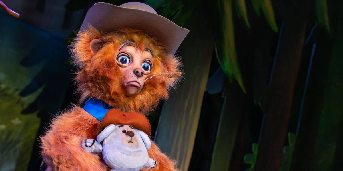 A fuzzy orange bear wearing a cowboy hat and blue scarf holds a small stuffed animal, looking surprised against lush green foliage—much like stumbling upon one of the Magic Kingdom underrated attractions.
