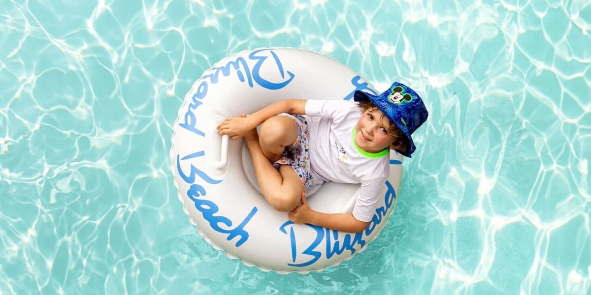 A young child wearing a blue hat, white shirt, and patterned swim trunks floats in a clear swimming pool on a white inner tube with blue writing at Disney Waterparks, smiling and looking up at the camera.