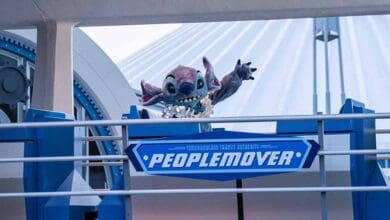 Stitch, a blue alien character, waves from behind a blue railing with a “PeopleMover” sign at a futuristic-themed amusement park attraction—one of the classic Disney World rides with short lines.