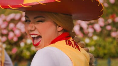 A person in a colorful costume and large hat is smiling widely and winking at the camera, enjoying Disney Employee Perks amid a blurred background of flowers and greenery.