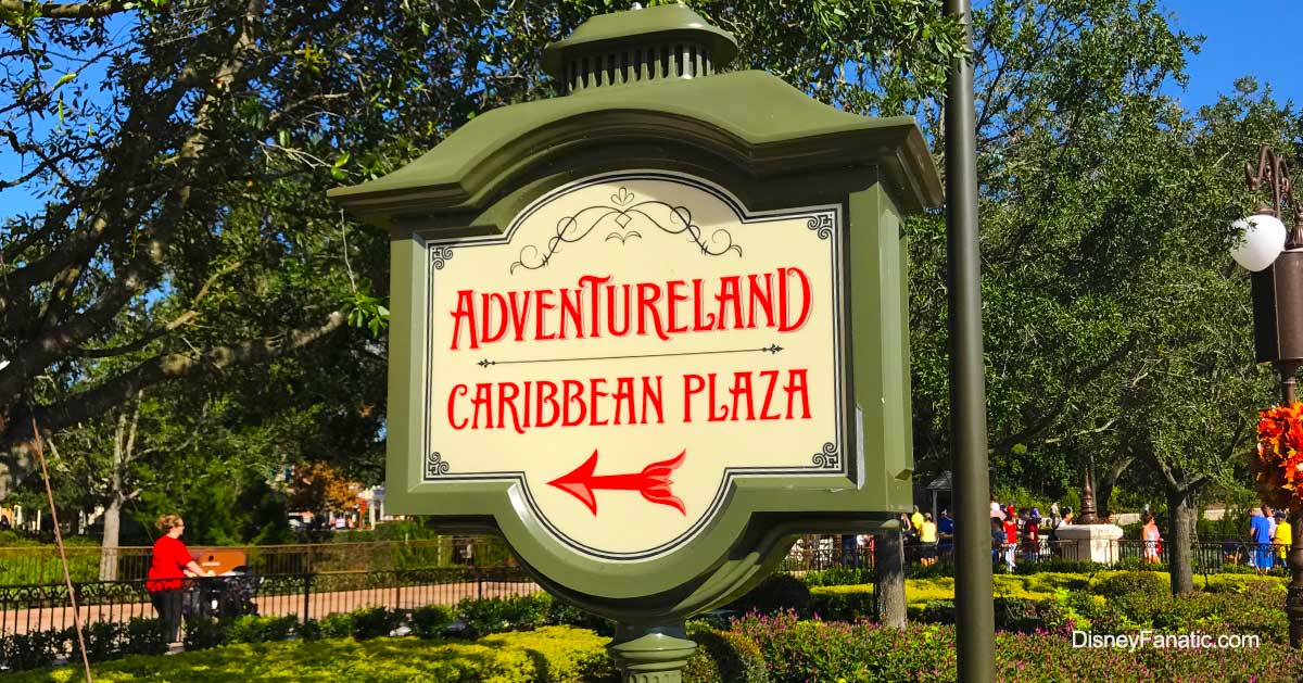 The image shows a sign with ornate detailing, pointing towards "Adventureland" and "Caribbean Plaza" in red lettering, with a red arrow. The sign is set against a green, park-like background.