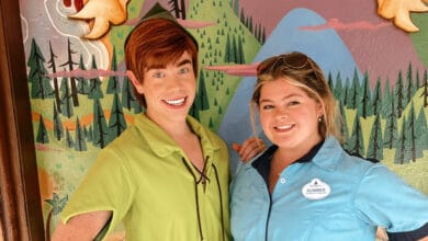 A person dressed as Peter Pan stands next to a smiling woman in a blue shirt and sunglasses, both posing in front of a colorful, painted forest-themed wall—showcasing the magic you can find with the right Disney Cast Member tips.
