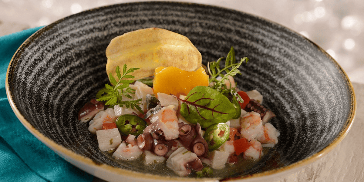 A bowl of seafood ceviche with shrimp, octopus, sliced green chili, diced tomatoes, herbs, a yellow sauce, and a plantain chip garnish—served on a dark patterned plate with a teal napkin—brings Disney World dining flair to your table.