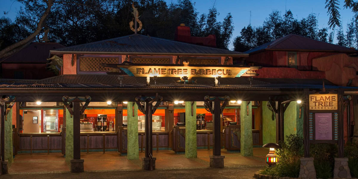 Flame Tree Barbecue restaurant at dusk, warmly lit with an inviting open counter, colorful pillars, and a large sign above the entrance—perfect for enjoying Healthy Dining at Disney Parks. Trees and a darkening sky are visible in the background.