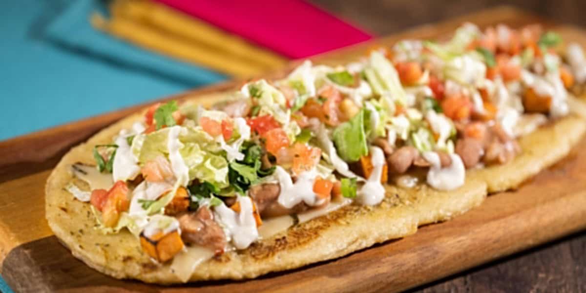 A Mexican sope topped with lettuce, diced tomatoes, beans, sour cream, and cheese—an ideal choice for Healthy Dining at Disney Parks—served on a wooden board with colorful napkins in the background.