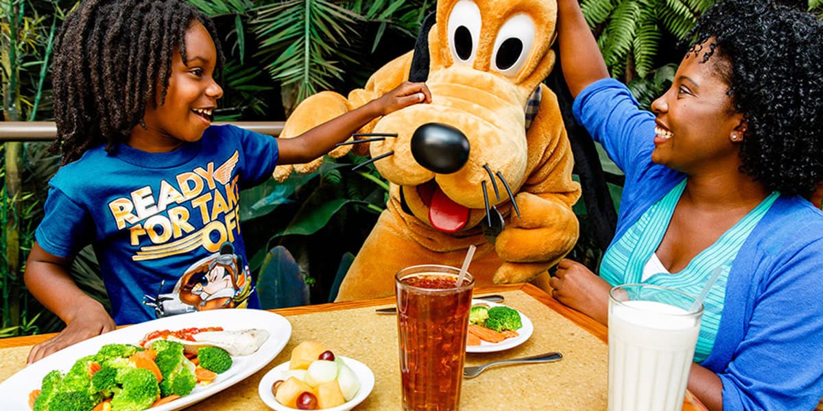 A smiling child and adult enjoy Healthy Dining at Disney Parks, sitting at a table with nutritious meals. Surrounded by lush greenery, they happily greet a person in a Pluto costume, both looking excited in this magical setting.