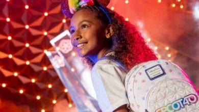 A young girl with curly hair wears colorful Mickey Mouse ears and a white backpack with an "Epcot 40" design. She smiles against a backdrop of bright lights and a futuristic building at a theme park.