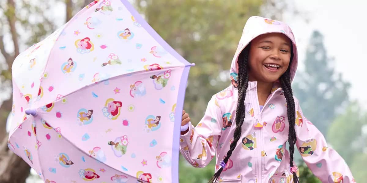 A smiling young girl holds a pastel-colored umbrella decorated with cartoon characters. She is wearing a matching hooded raincoat with similar designs. The background is an outdoor setting with trees.