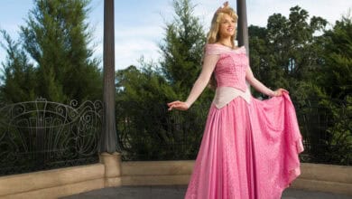 Aurora in a bright pink ball gown and crown stands smiling on a gazebo with iron railings, surrounded by greenery and trees under a blue sky.