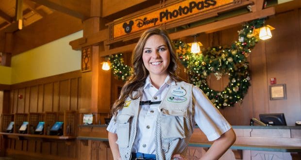 A Disney PhotoPass photographer beams in festive attire by a holiday wreath, capturing memories during modified park operations.