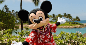 Mickey Mouse wearing a red Hawaiian shirt and a purple lei poses cheerfully with arms wide open in front of tropical plants, palm trees, and a lagoon under a blue sky.