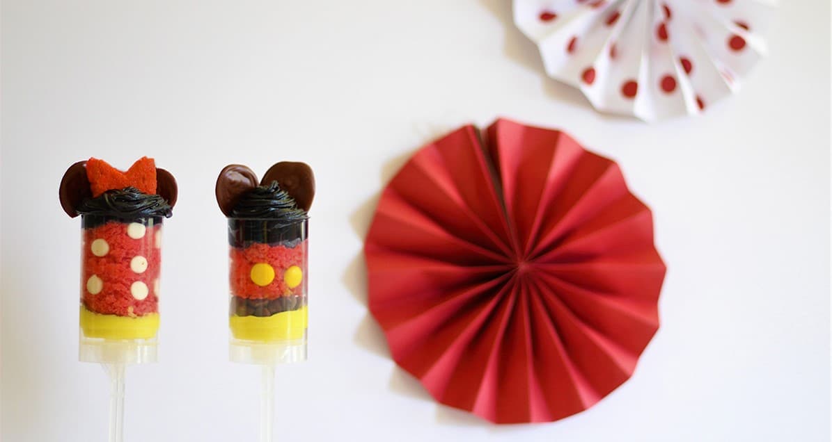 Minnie Cake Push Pops with red and yellow layers are displayed at a Disney park, surrounded by themed paper decorations.