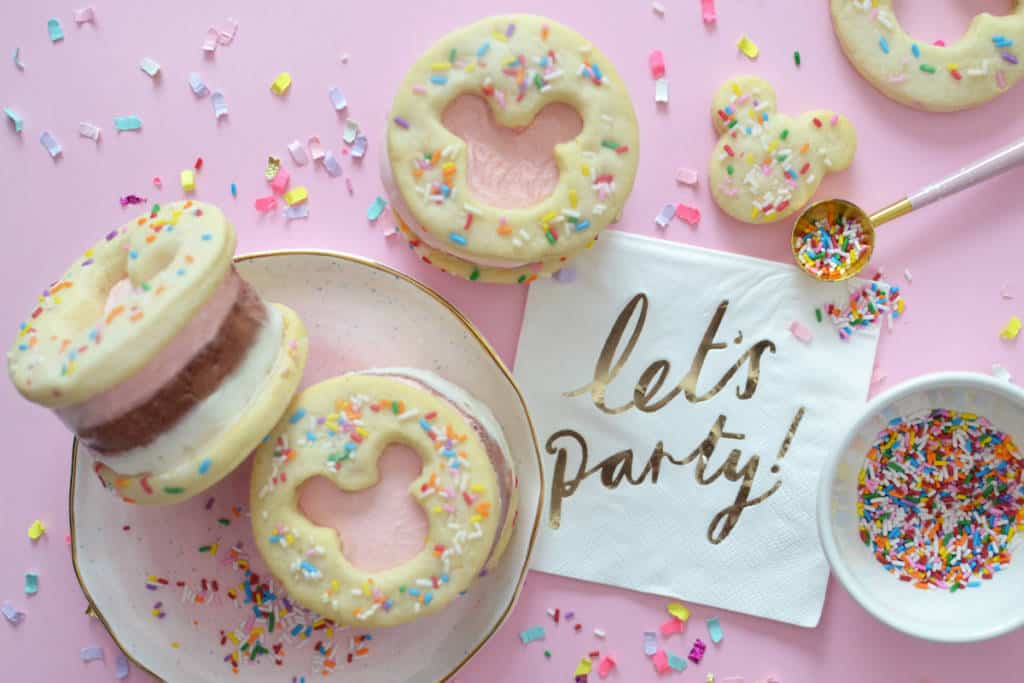 Disney-inspired ice cream sandwiches with Hidden Mickey cutouts and sprinkles, on a pink background beside a “let’s party!” napkin.