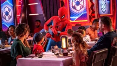 Spider-Man interacts with children at a themed restaurant aboard the Disney Cruise Line, surrounded by families enjoying their meals. The vibrant setting features red and blue superhero decor and futuristic lighting.