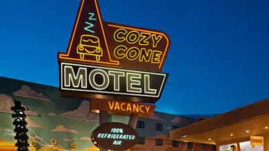 A brightly lit neon sign reading “Cozy Cone Motel” with a cartoon car and cone design glows against a dusk sky, advertising vacancy and “100% Refrigerated Air”—a whimsical stop featured in the Disney World Resort Guide. A building and mural are visible behind it.