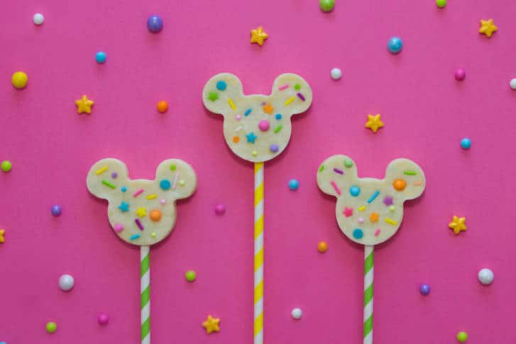 Whimsical Mickey Mouse-shaped lollipops with colorful sprinkles, inspired by Disney treats, on a playful pink starry backdrop.