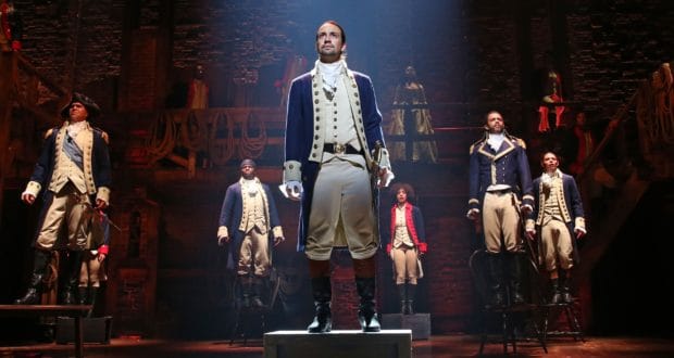 Hamilton cast in iconic colonial attire delivers a dynamic stage show under vibrant lights, captivating the Broadway audience.