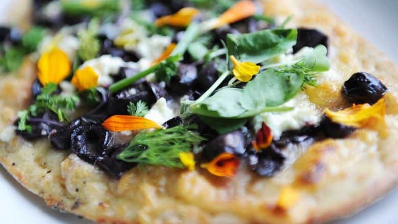 Jiko’s millet flatbread from Disney’s Animal Kingdom, topped with olives, greens, cheese, and petals on a white plate.