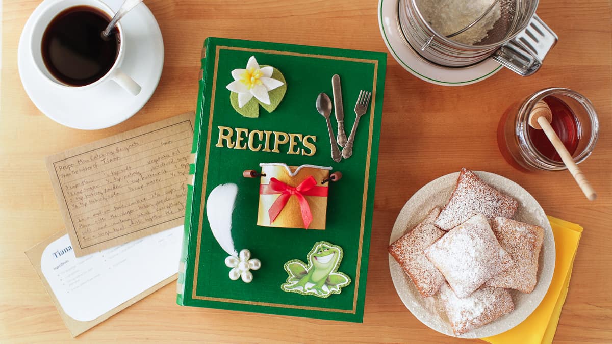 A Tiana-themed green recipe book at Disney sits with coffee, beignets, honey, and a recipe card—ideal for Disney DIY fun.