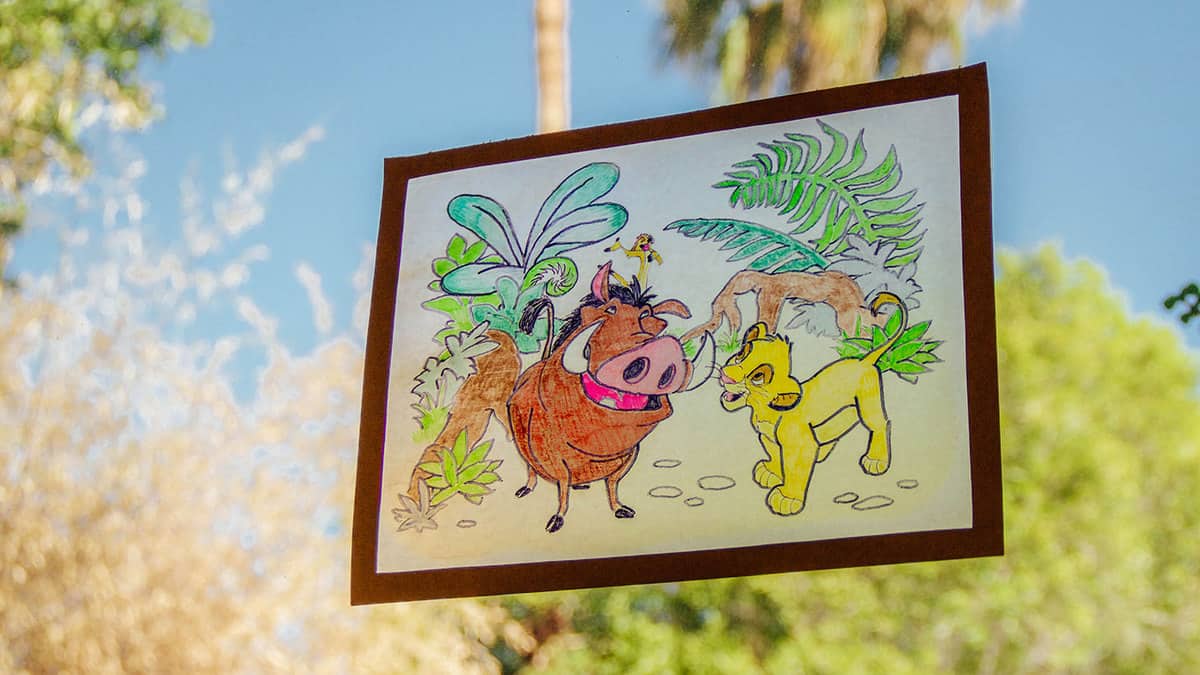 A colored Disney page of Pumbaa and Simba shines on a window, creating a sunny, magical Lion King vibe for fans.