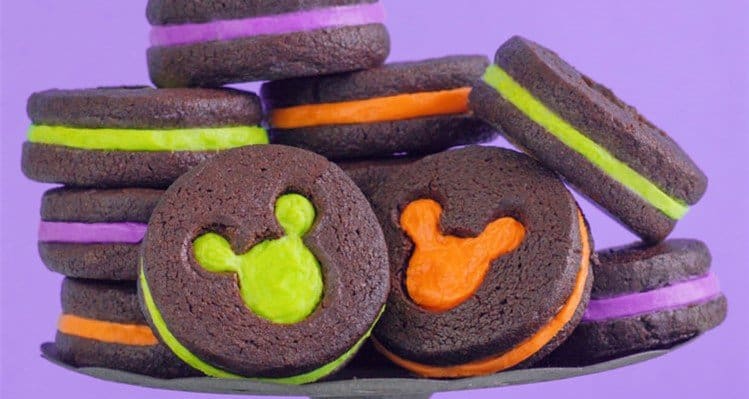 Halloween-themed Mickey Mouse chocolate sandwich cookies with vibrant fillings, stacked in front of a purple Disney Park backdrop.