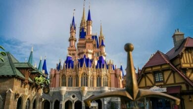 A view of Cinderella’s Castle with blue spires at Disney World, surrounded by whimsical buildings and an oversized sword in the foreground, under a clear blue sky.
